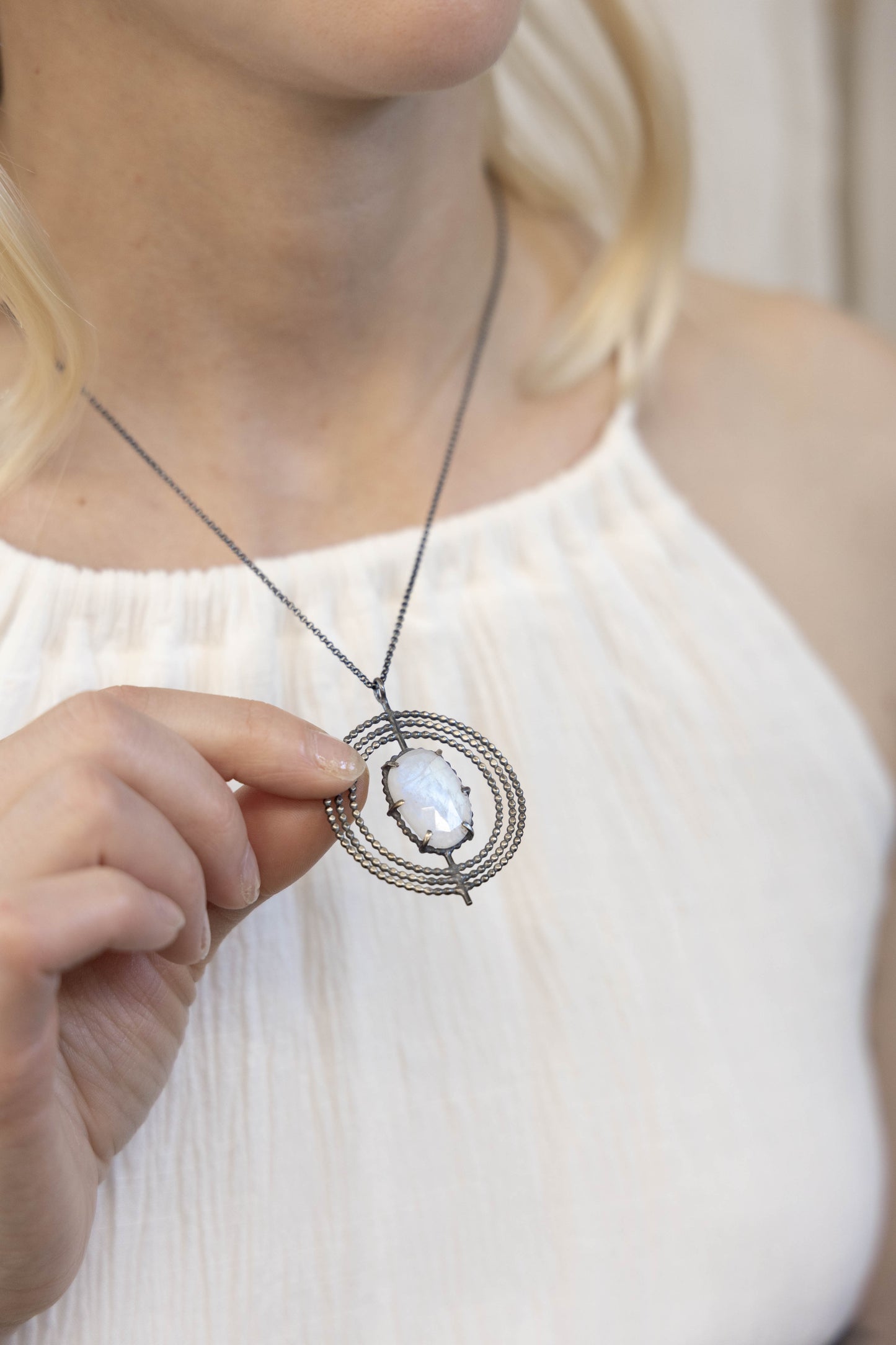 A necklace with a central moonstone surrounded by a circular silver frame with intricate details, on a model, by Alice Scott