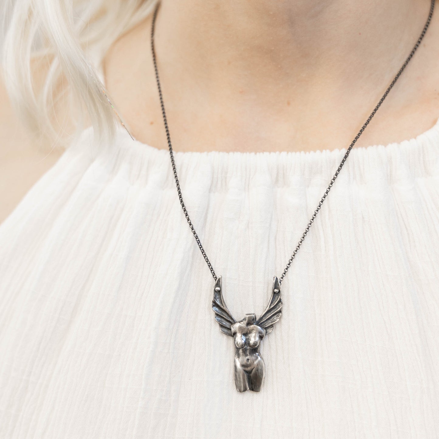 A silver necklace with a detailed pendant representing the Winged Venus figure, suspended on a black patina chain on a model neck, by Alice Scott