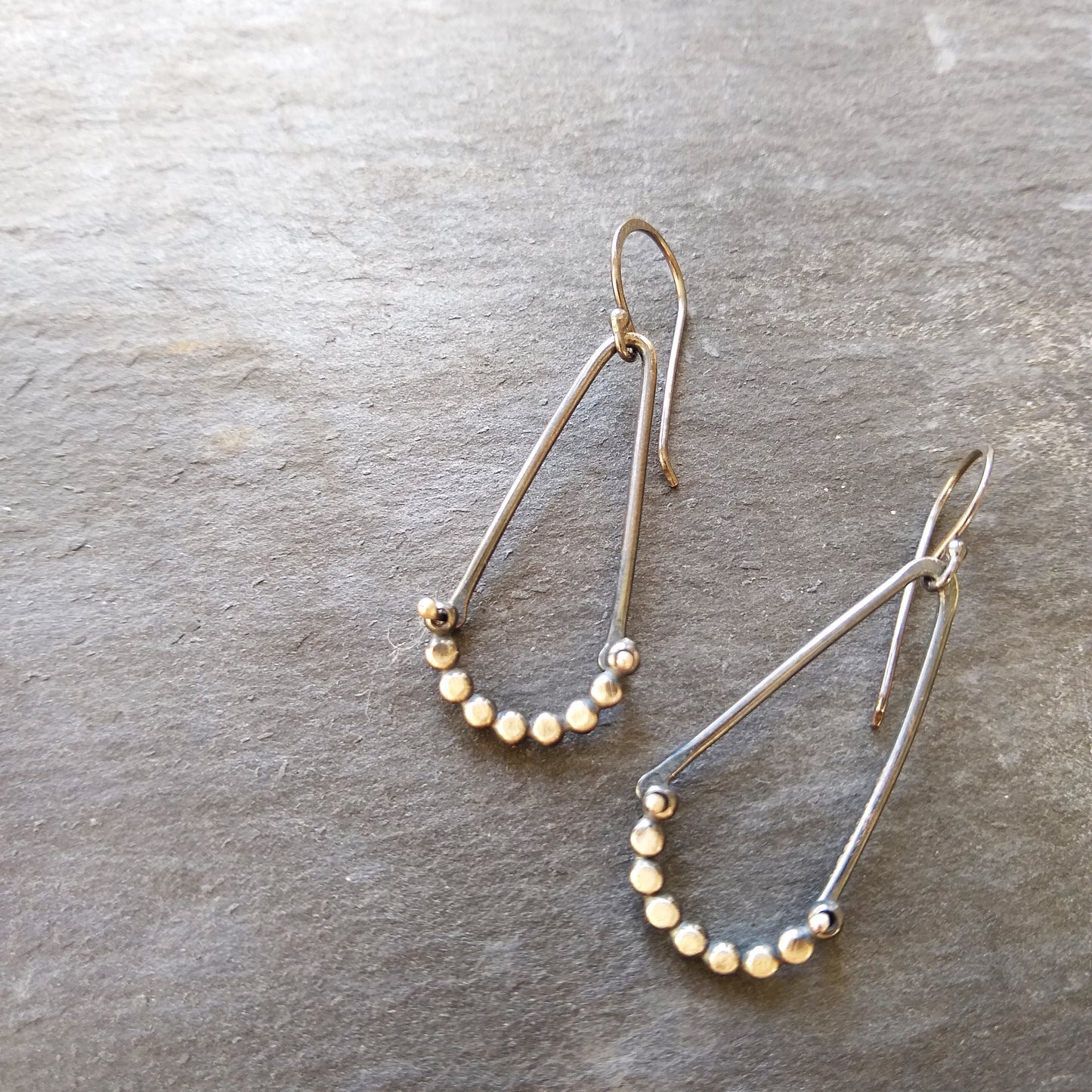 A pair of silver earrings with a hammered dot pattern attached to oxidized silver angle shapes, displayed against a textured grey background by Alice Scott