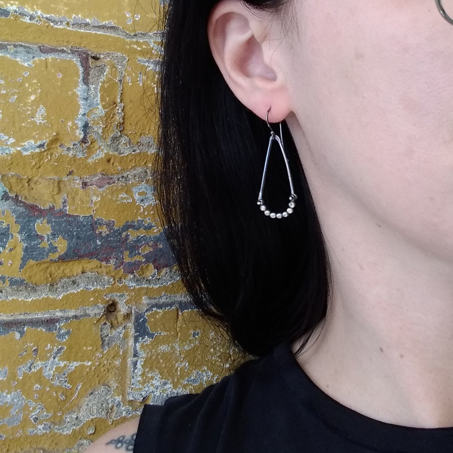 A pair of silver earrings with a hammered dot pattern attached to oxidized silver angle shapes, displayed against a model ear by Alice Scott