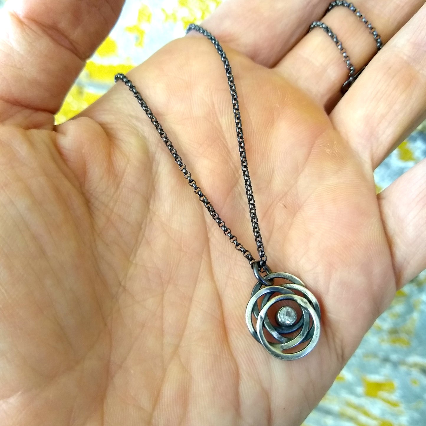 A sterling silver necklace with a black patina featuring a flowing circle pendant containing a glittering white topaz, displayed in a hand. The necklace is by Alice Scott
