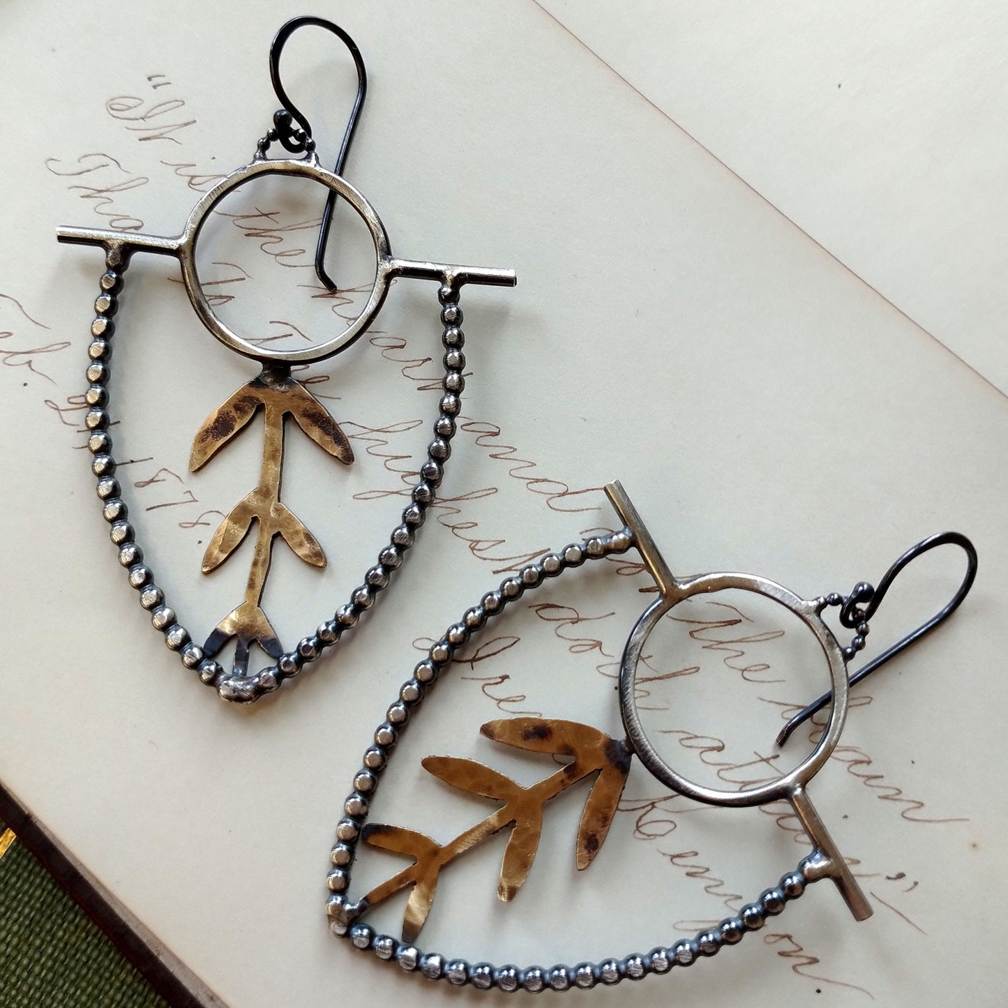 A pair of earrings with brass sprouts and sterling silver earwires and dots with a black patina, displayed on a piece of paper by Alice Scott