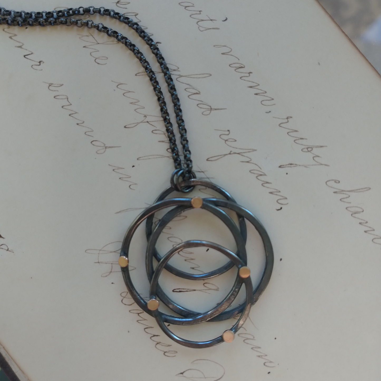 A silver necklace with a hammered galaxy pendant featuring gold dots, displayed on a piece of paper with writing, by Alice Scott