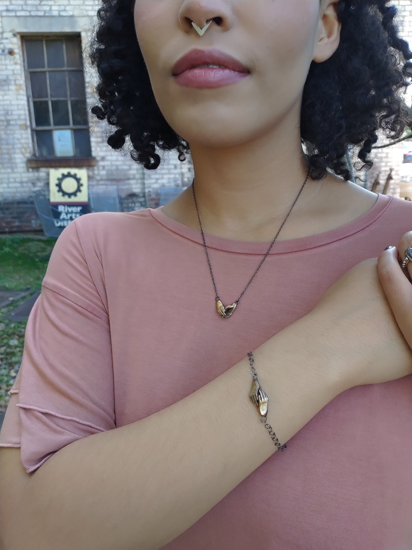 A bronze bracelet featuring a pair of hands holding each other, linked together, with a silver chain and black patina by Alice Scott on model background