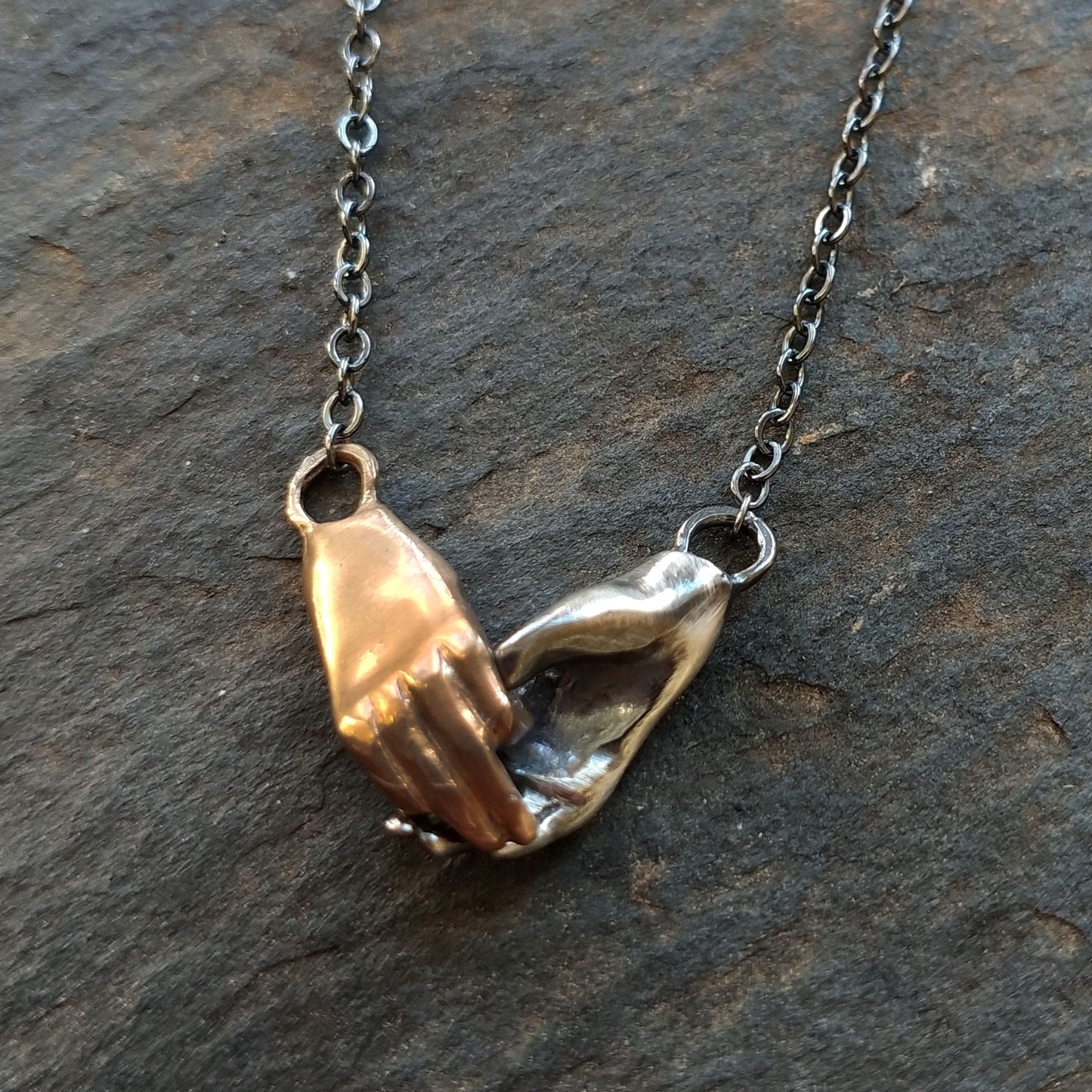 A bronze and silver necklace in the shape of two hands holding each other, displayed against a stone background, by Alice Scott