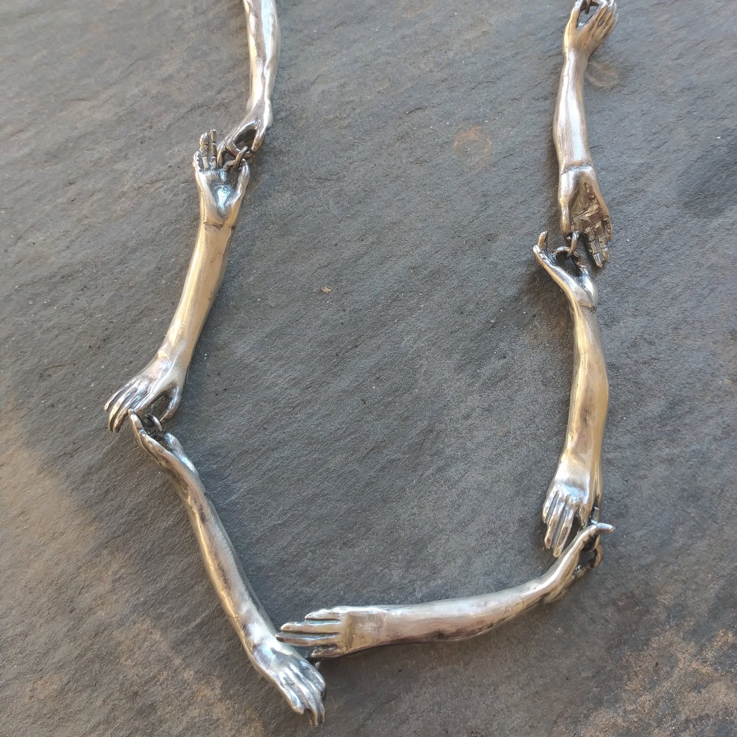 A silver necklace with a unique design featuring hand-shaped links connected by silver rings on a silver chain, by Alice Scott, on gray background.