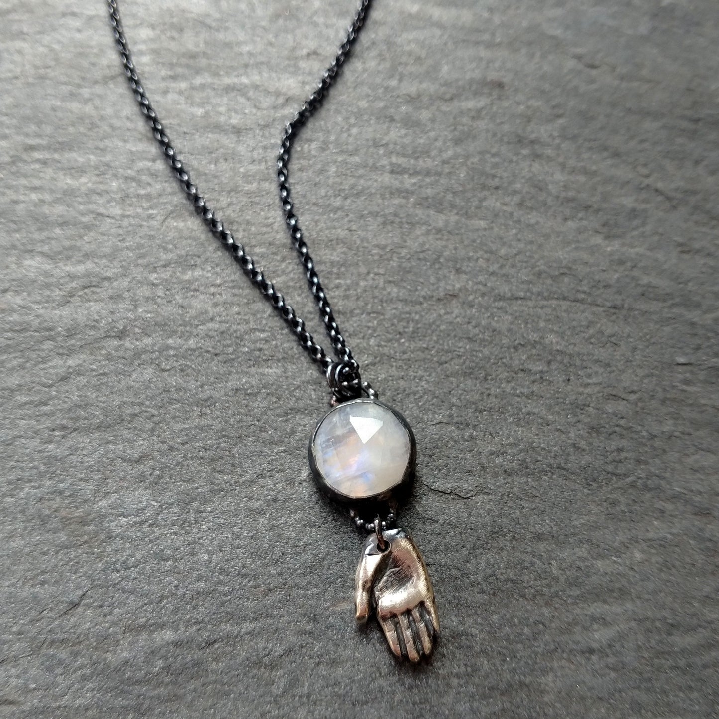 A silver necklace with a rainbow moonstone pendant and a hand talisman charm on a dark textured background, by Alice Scott