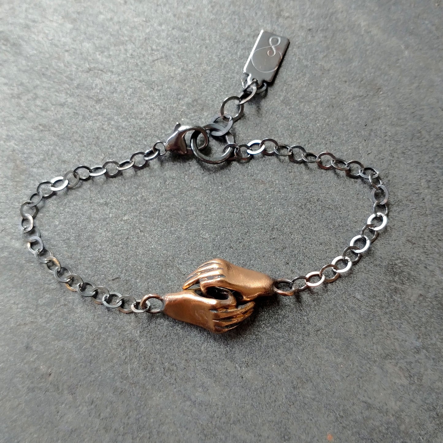 A bronze bracelet featuring a pair of hands holding each other, linked together, with a silver chain and black patina by Alice Scott on grey background