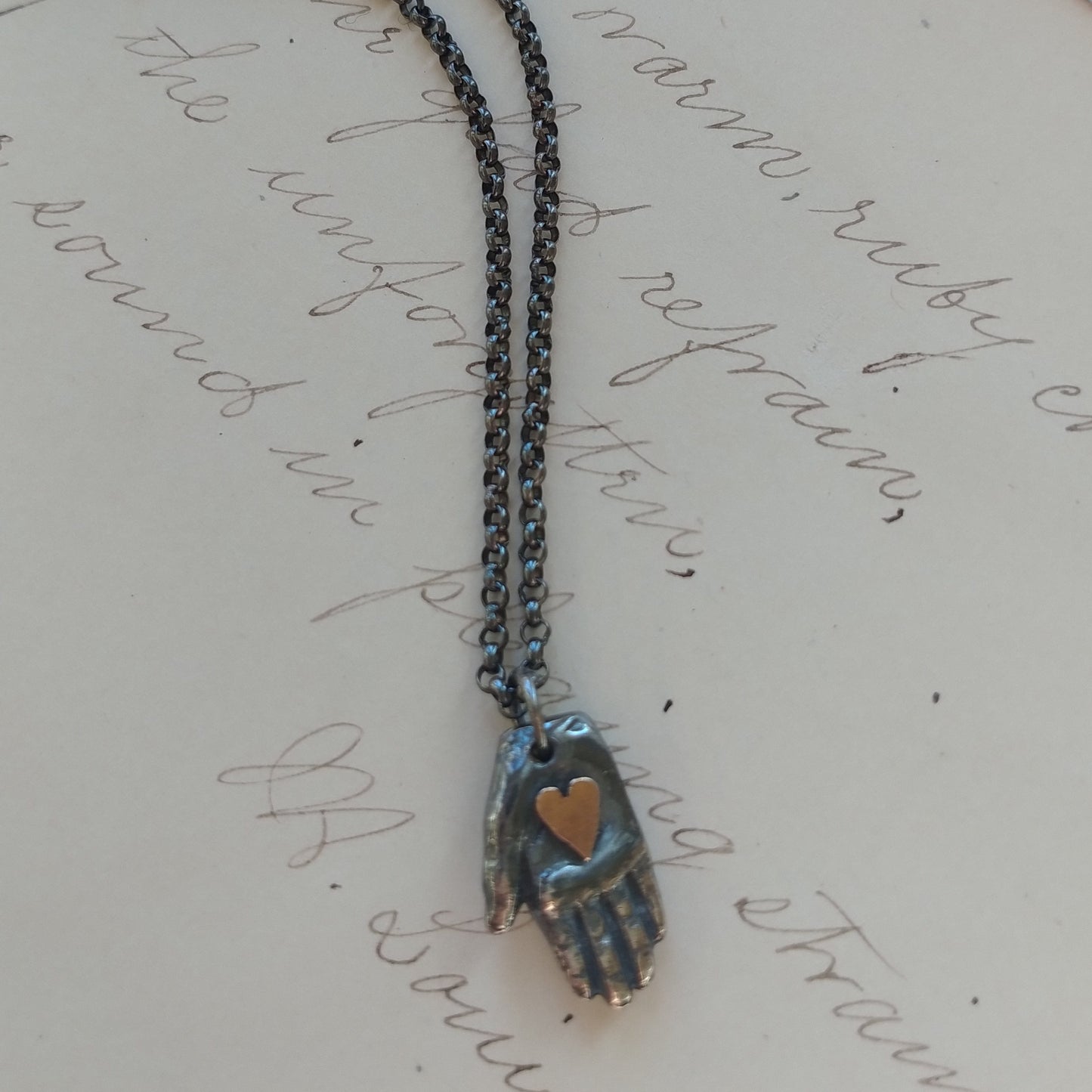 tiny hand necklace with golden heart pendant with silver chain on written background by Alice Scott