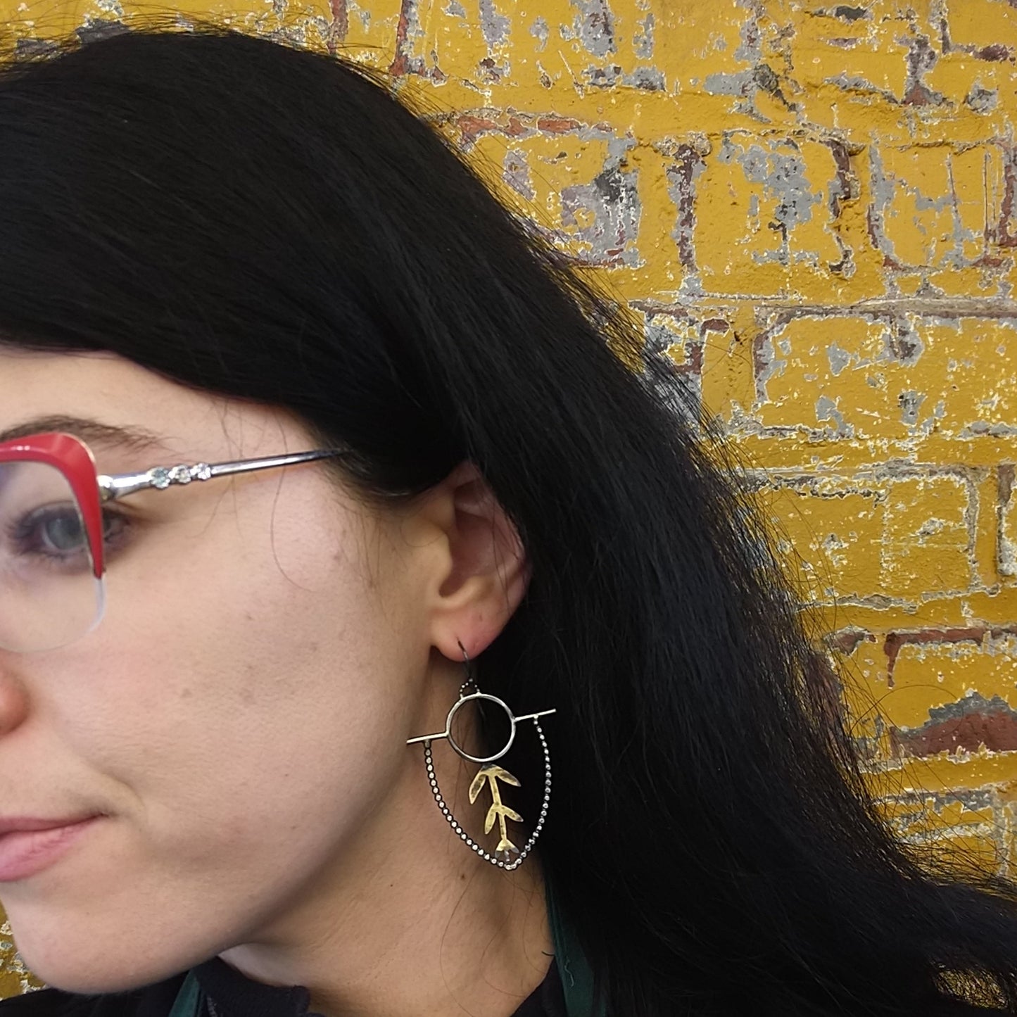 A pair of earrings with brass sprouts and sterling silver earwires and dots with a black patina, displayed on a model ear, by Alice Scott
