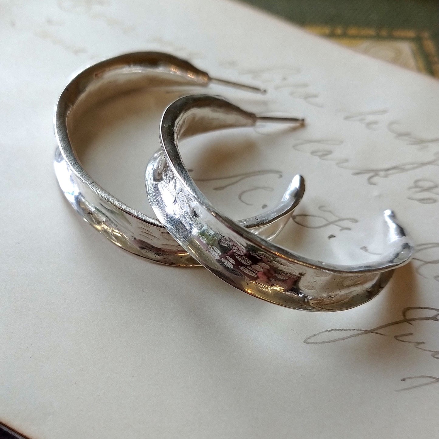 A pair of sterling silver hammered hoop earrings resting on a piece of paper with writing, by Alice Scott