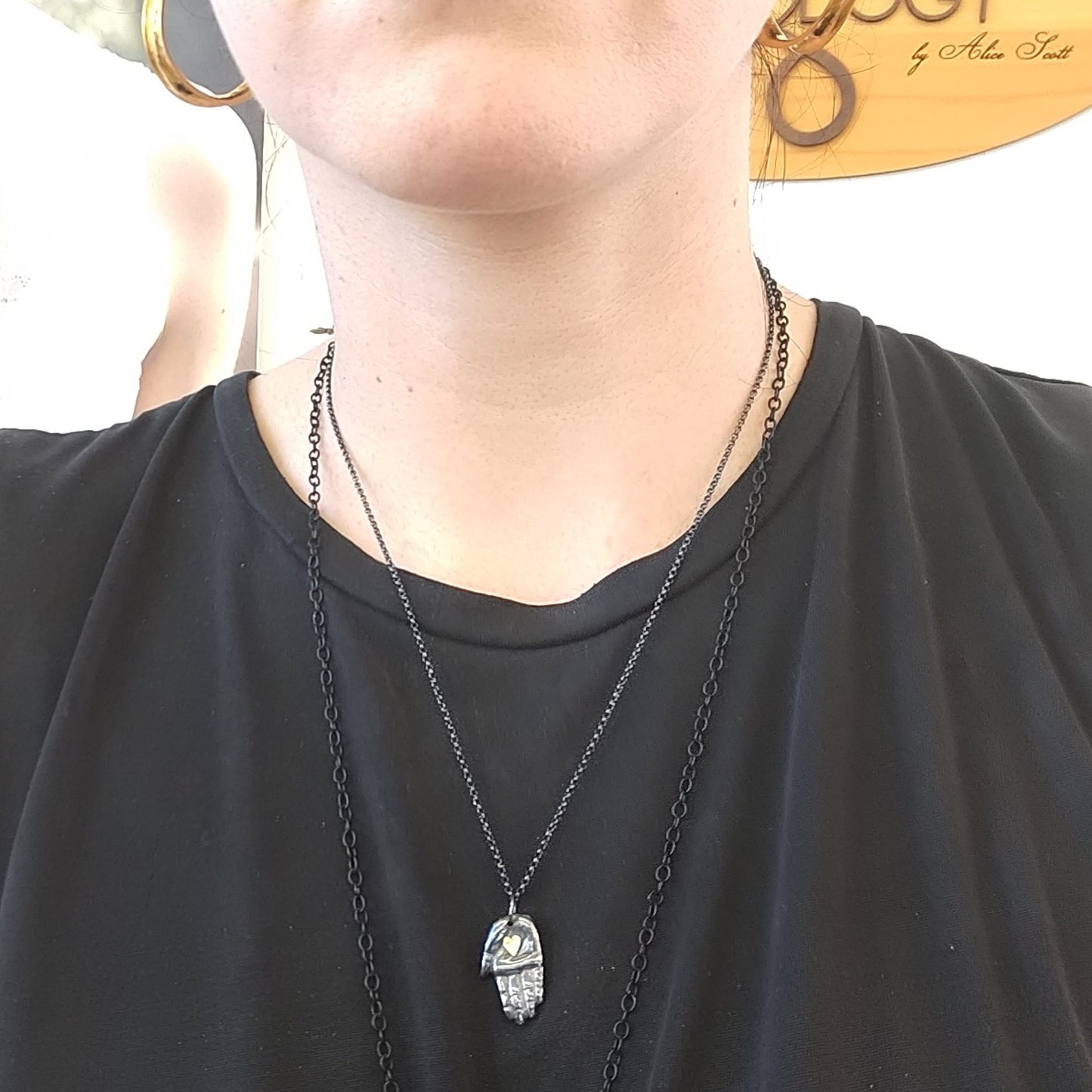 A silver necklace with a handmade patina finish featuring a hand with a gold heart pendant on a chain, displayed on a model neck, by Alice Scott.
