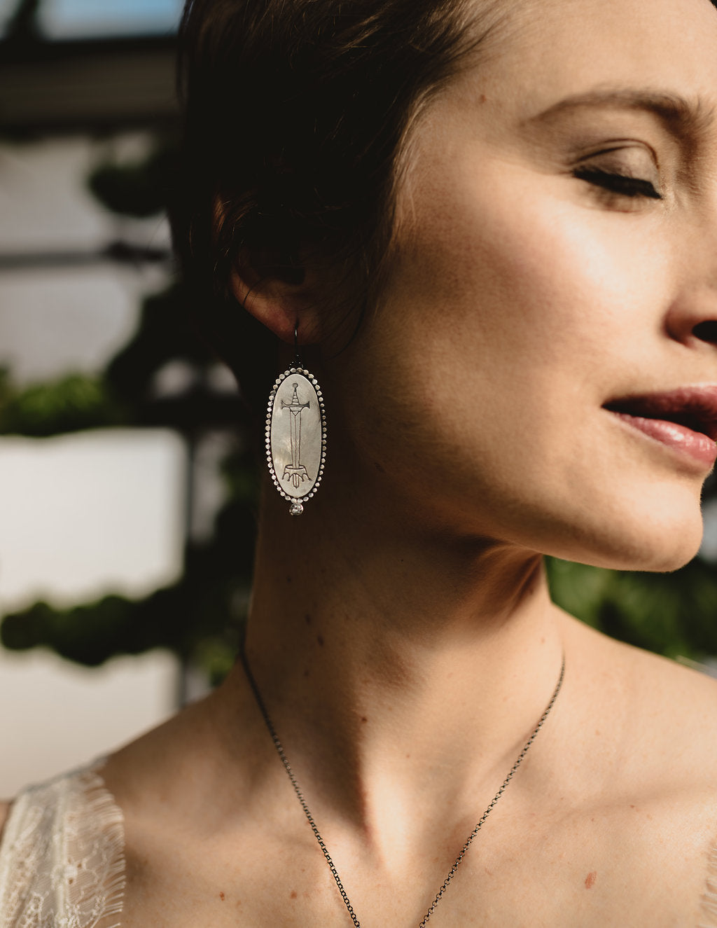 Silver Oval Earrings with sword engraving, surrounded by wilver dots and a white topaz at the bottom, by Alice Scott, displayed on a model ear