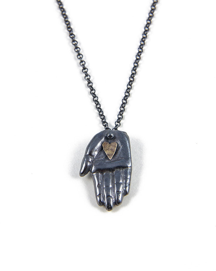 A silver necklace with a handmade patina finish featuring a hand with a gold heart pendant on a chain, displayed on a white background, by Alice Scott.