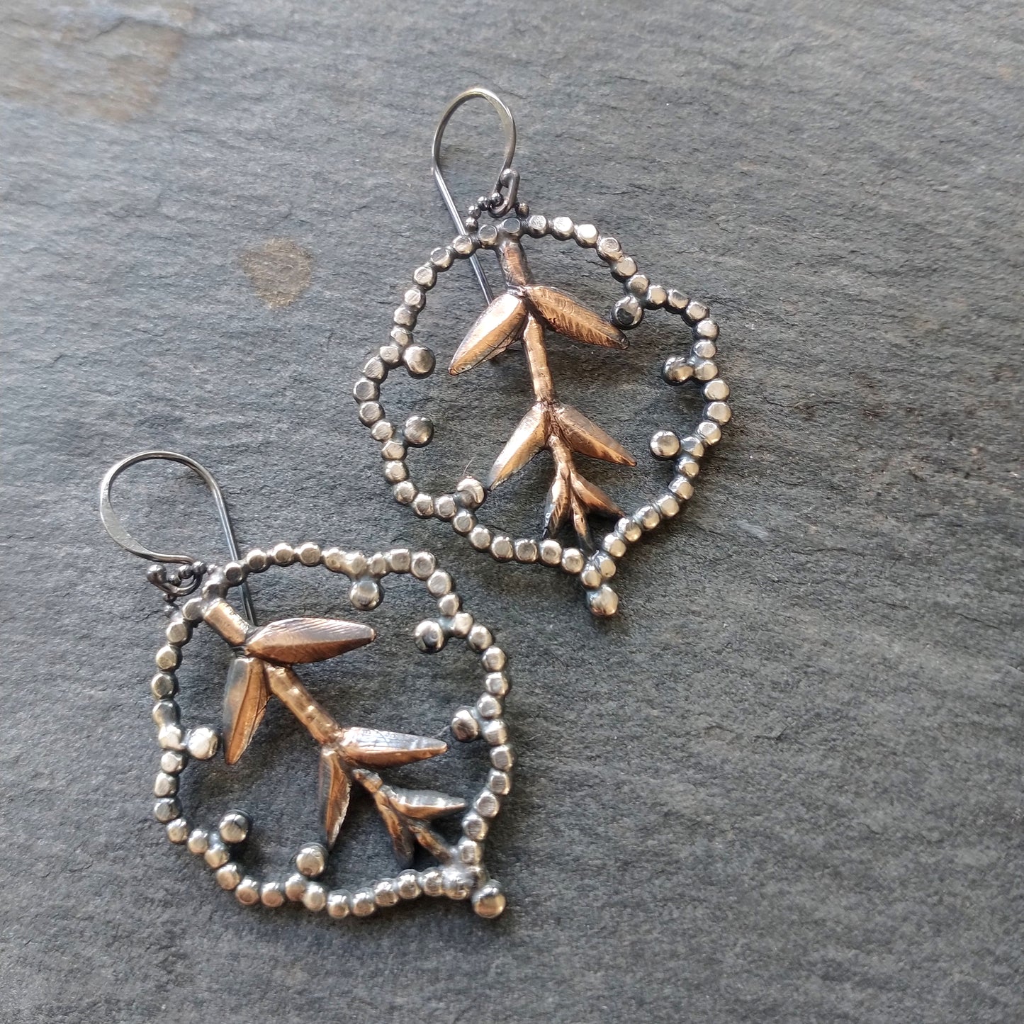 A pair of earrings featuring a lacy cloud design with bronze roots and silver accents on a slate background by Alice Scott.