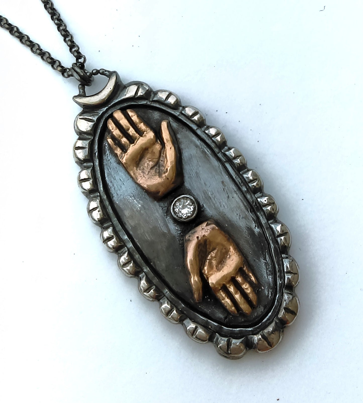 Close-up of a double hand cameo necklace with bronze hands and a sterling silver cameos background, featuring a topaz gemstone between the hands, by Alice Scott, on a white background
