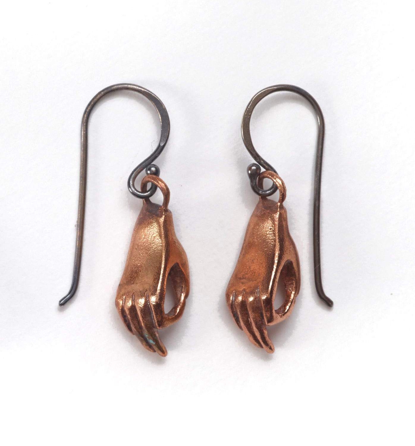 A pair of bronze earrings in the shape of hands performing a mudra, attached to oxidized silver wire hooks, set against a white background by Alice Scott