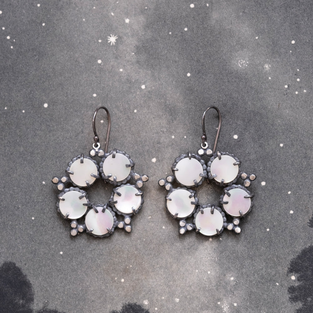 A pair of silver, mother of pearl earrings with a star and constellation design, displayed against a dark background speckled with stars by Alice Scott