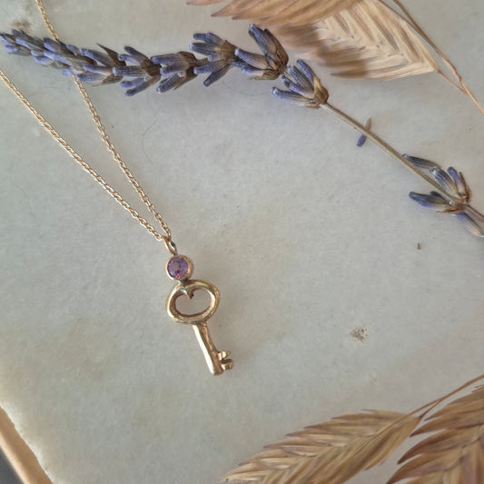Gold key-shaped pendant with a purple gemstone on a chain, set against a decorative background with leaves and flowers.