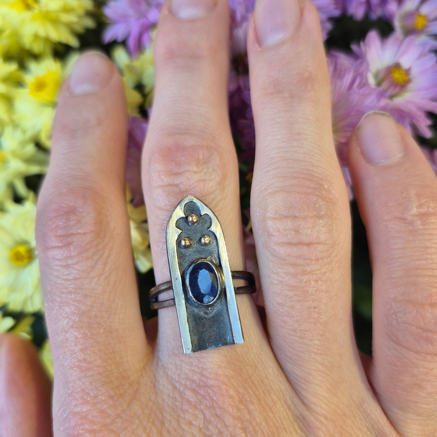 Hand wearing a silver ring with a blue sapphire with gold dots, by Alice Scott, against a floral background
