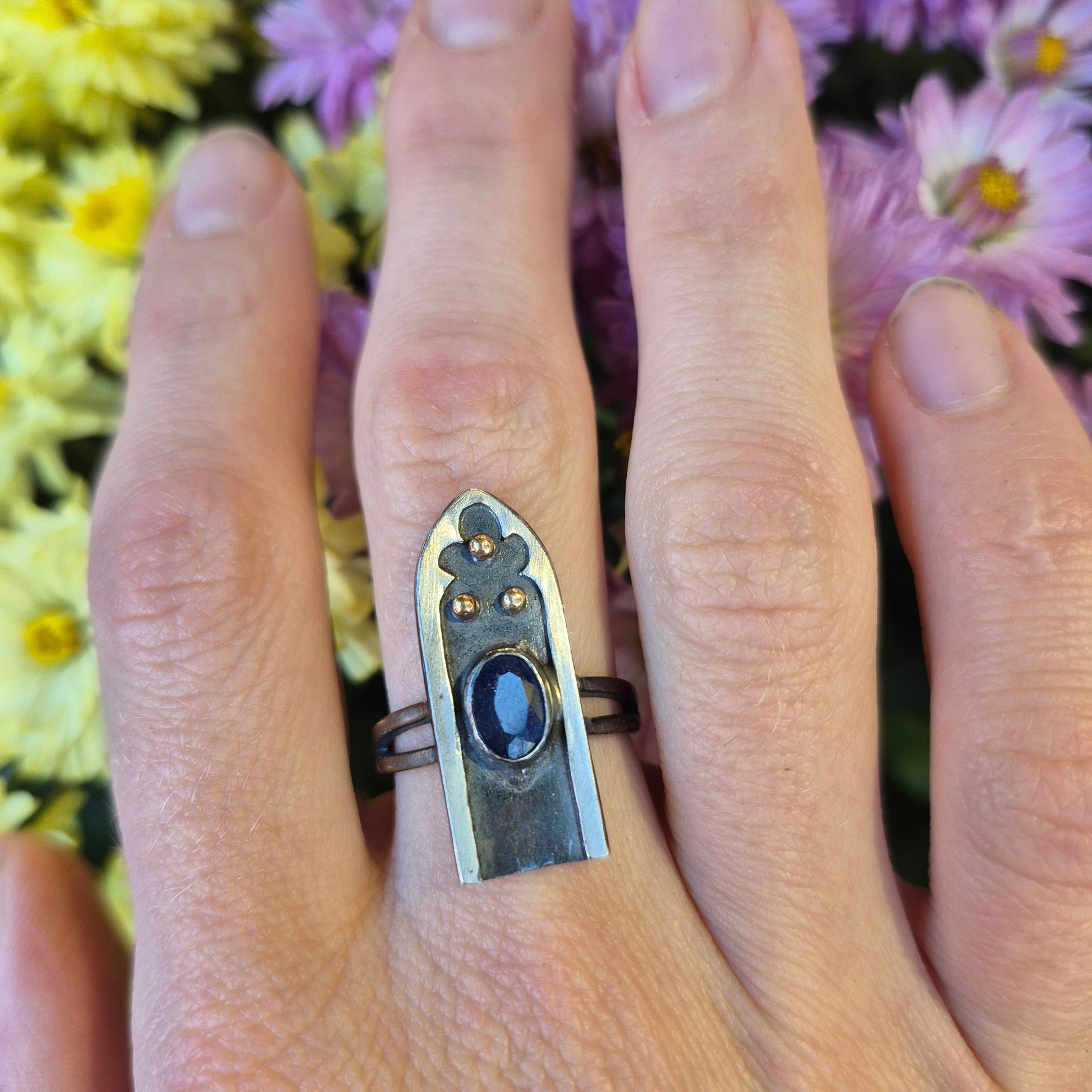 Hand wearing a silver ring with a blue sapphire with gold dots, by Alice Scott, against a floral background