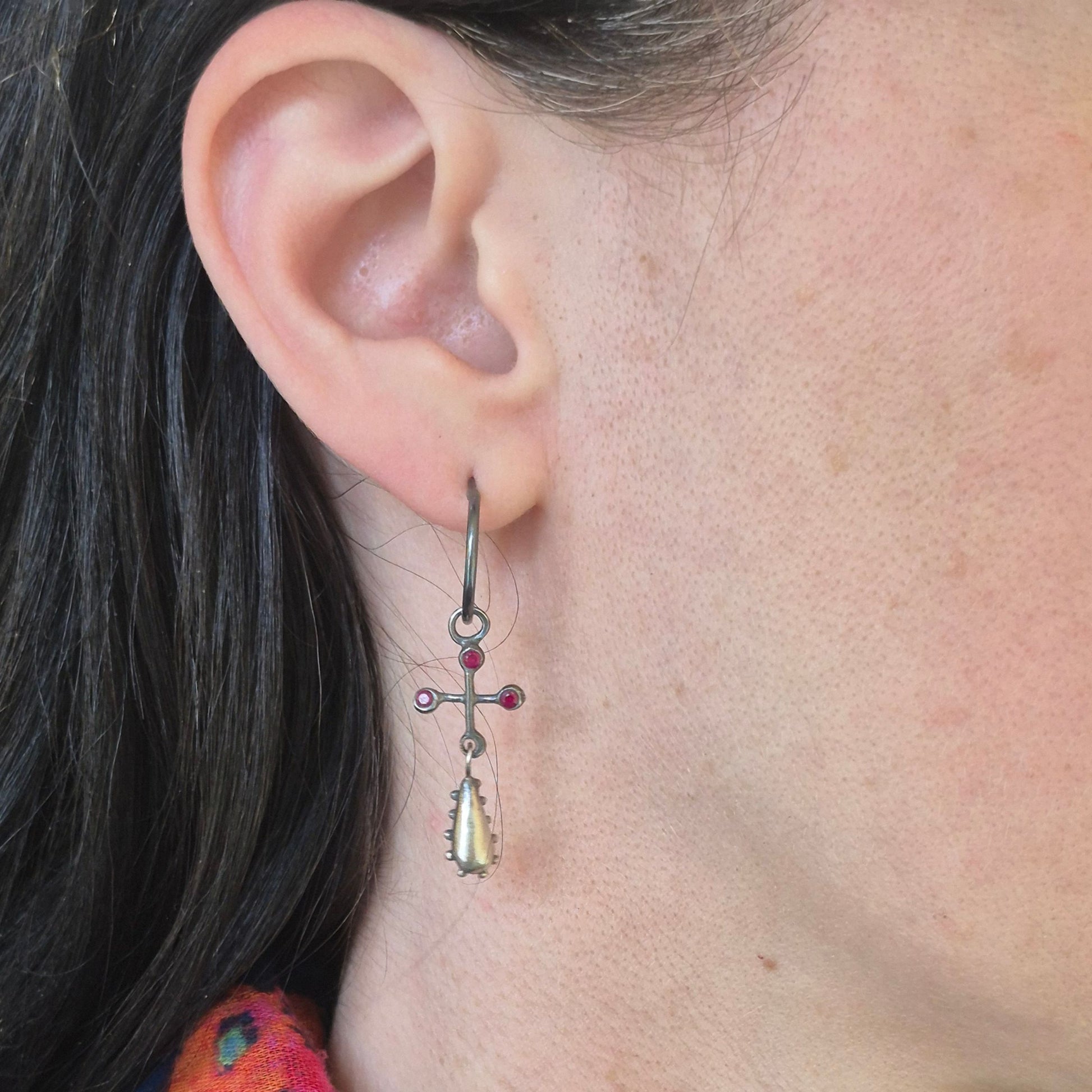 Close-up of an ear wearing a dangling earring with a cross design and rubies by Alice Scott