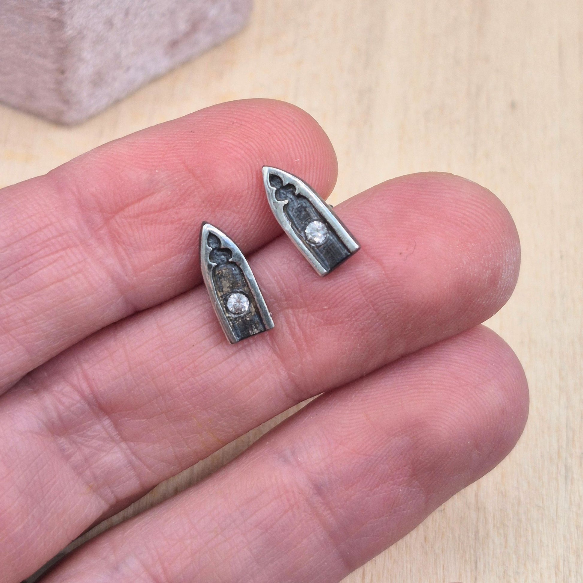 Silver gothic arch tiny post earrings with white sapphire gemstones held between fingers on a light wooden surface by Alice Scott