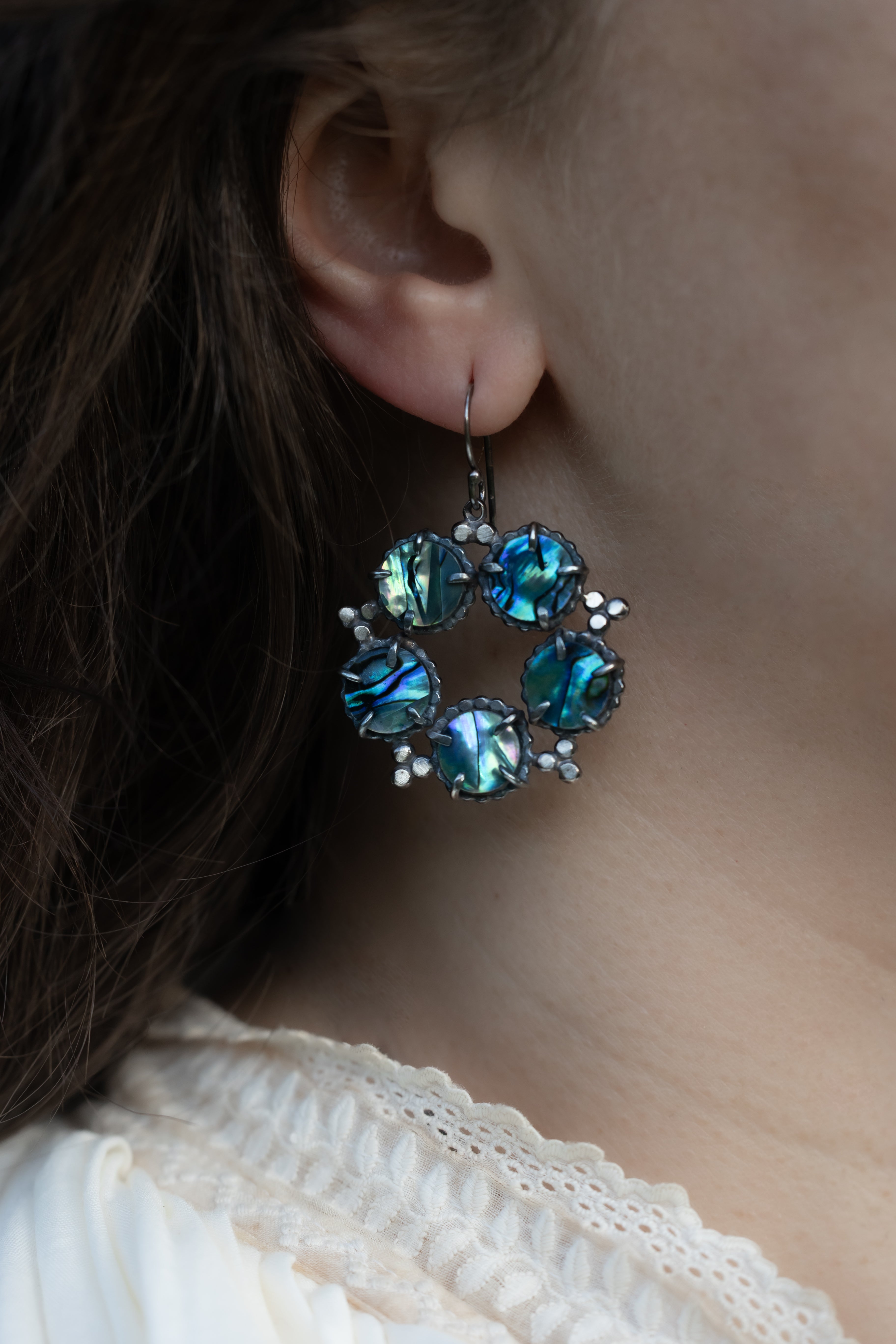 Close-up of a person wearing blue abalone shell earrings with silver on a blurred background by Alice Scott