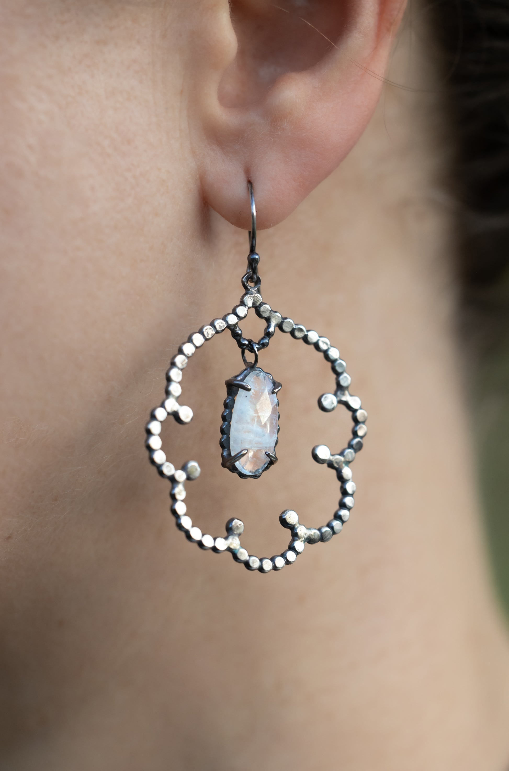 Close-up of an ornate silver earring with a central moonstone gemstone worn by a person, by Alice Scott