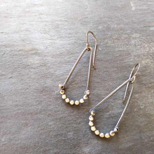 A pair of silver earrings with a hammered dot pattern attached to oxidized silver angle shapes, displayed against a textured grey background by Alice Scott