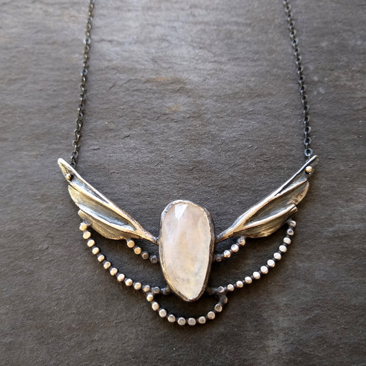 A silver necklace with a moonstone wing pendant and small silver accents on a slate background by Alice Scott
