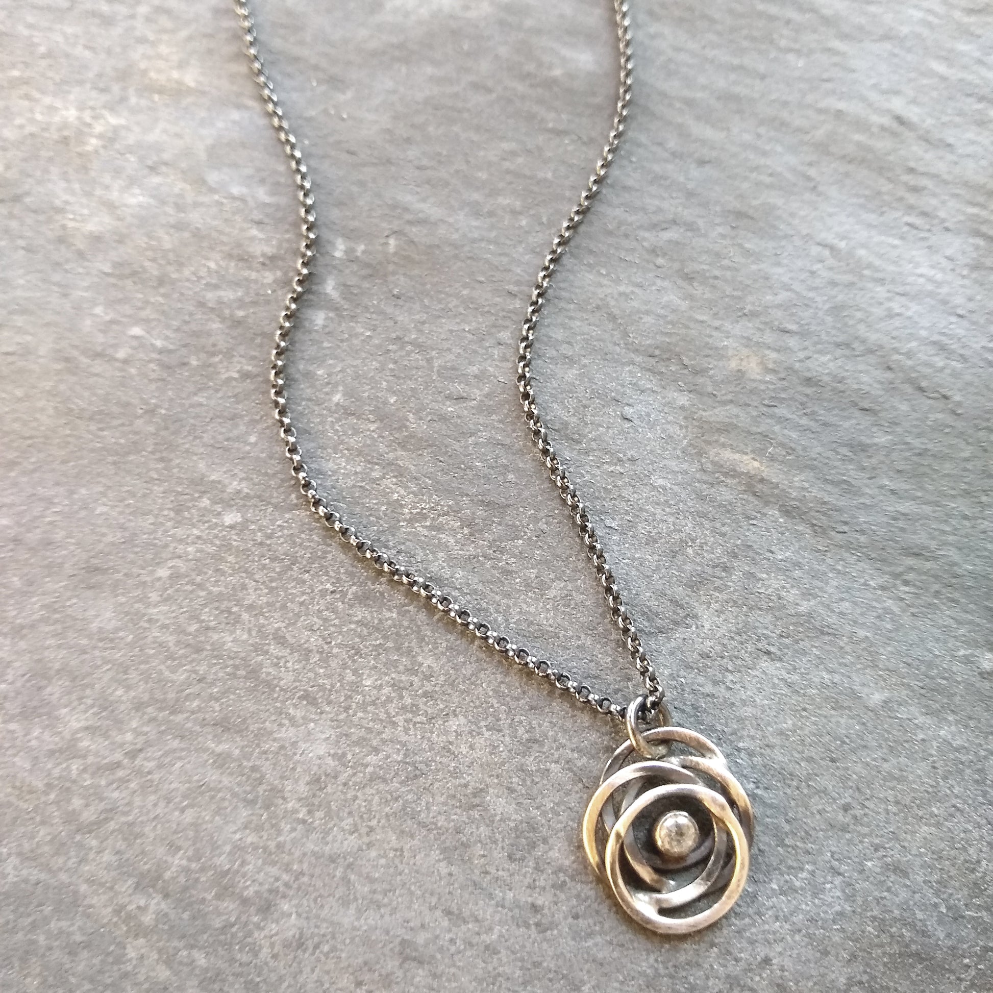 A sterling silver necklace with a black patina featuring a flowing circle pendant containing a glittering white topaz, displayed on a textured slate background by Alice Scott