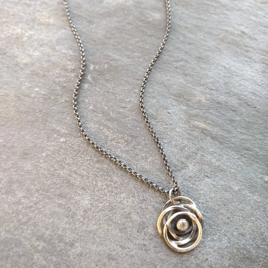 A sterling silver necklace with a black patina featuring a flowing circle pendant containing a glittering white topaz, displayed on a textured slate background by Alice Scott