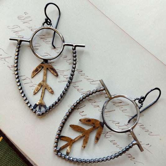 A pair of earrings with brass sprouts and sterling silver earwires and dots with a black patina, displayed on a piece of paper by Alice Scott