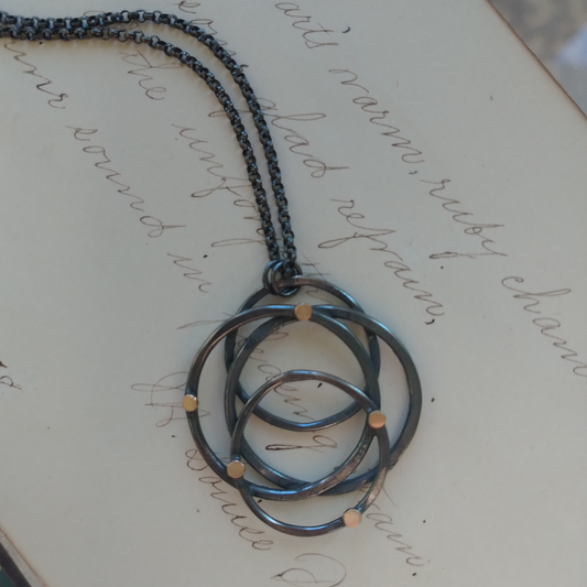 A silver necklace with a hammered galaxy pendant featuring gold dots, displayed on a piece of paper with writing, by Alice Scott