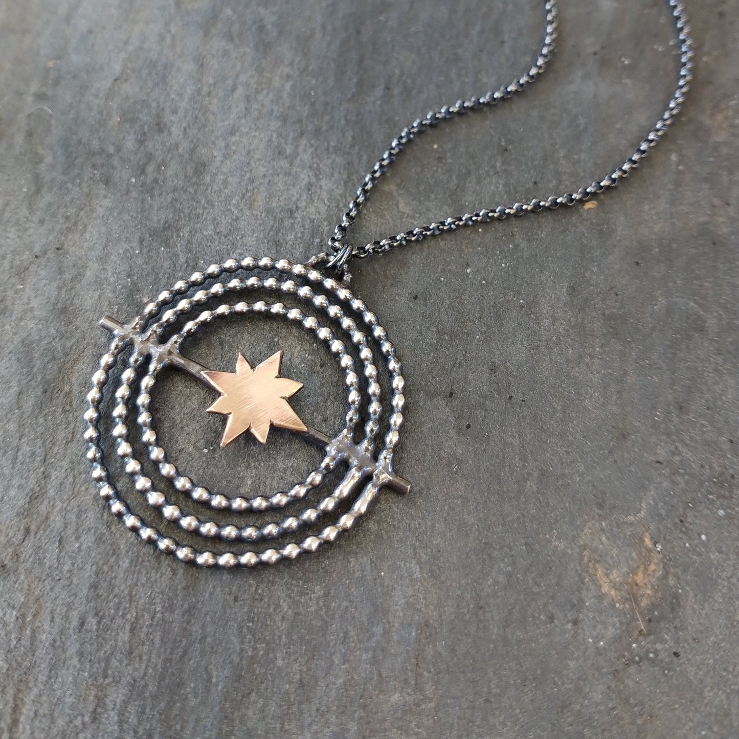 A sterling silver necklace with a gold fill star in the center, surrounded by a circular design, placed on a stone surface, by Alice Scott