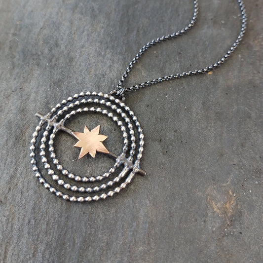 A sterling silver necklace with a gold fill star in the center, surrounded by a circular design, placed on a stone surface, by Alice Scott