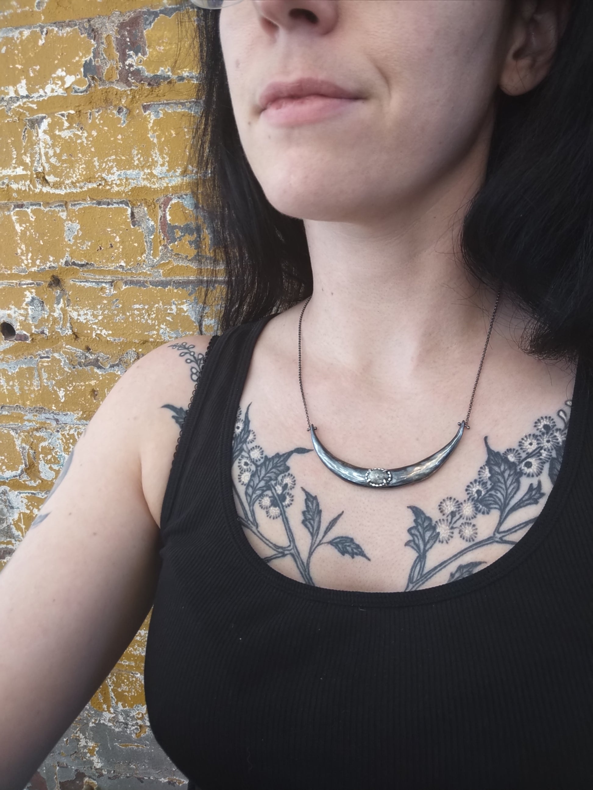 A silver crescent moon necklace with a black patina finish and a central aquamarine pendant, displayed against a mode, by Alice Scott