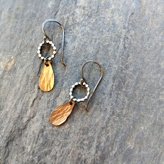A pair of handmade dangle earrings with gold drops and sterling silver components, displayed on a textured surface, by Alice Scott