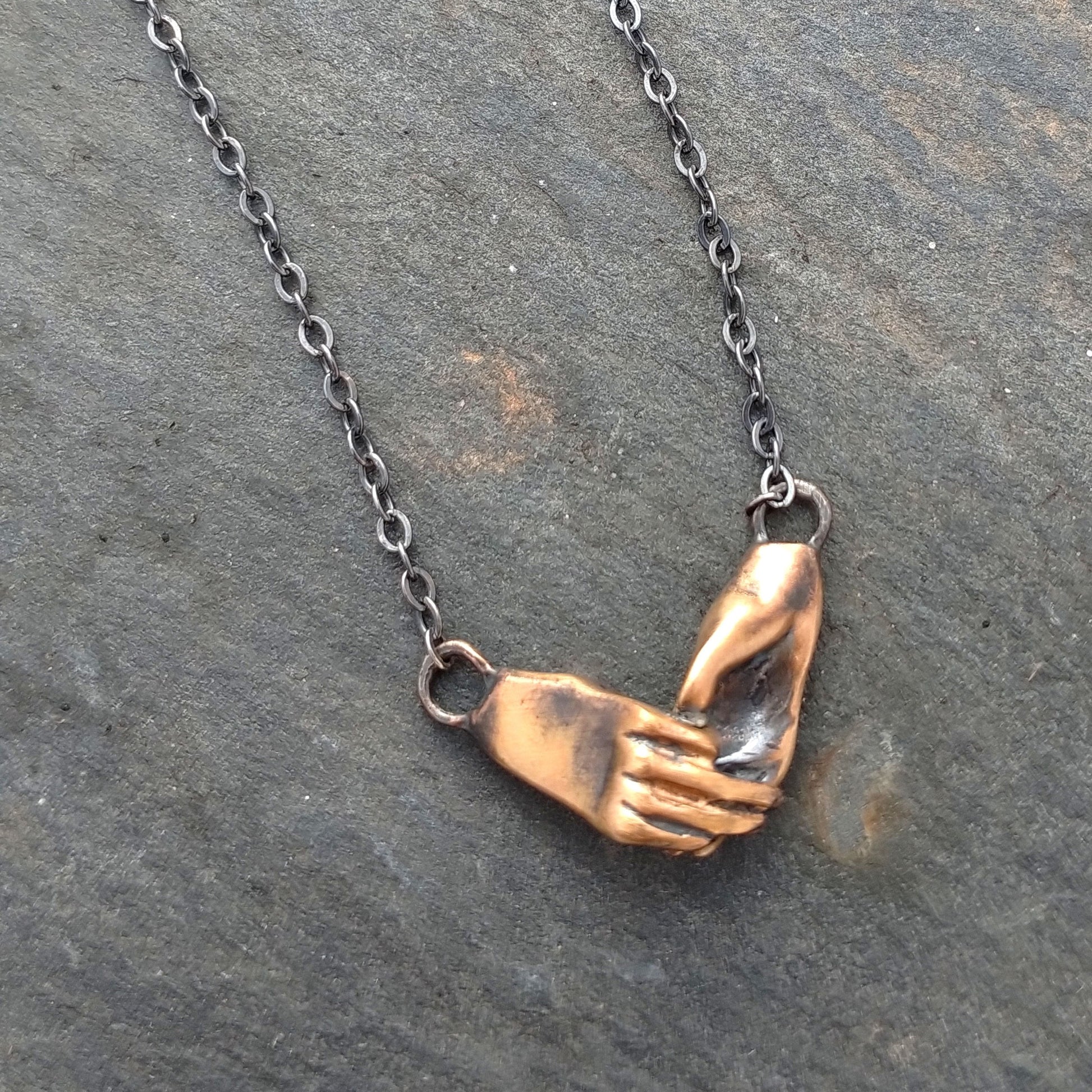 A bronze necklace featuring two hands holding each other, attached to a sterling silver chain with a sterling clasp, by Alice Scott on a grey background