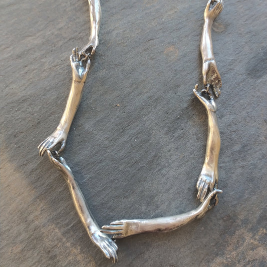 A silver necklace with a unique design featuring hand-shaped links connected by silver rings on a silver chain, by Alice Scott, on gray background.