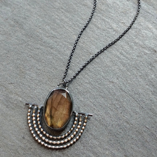 A sterling silver necklace with a black patina featuring an outstretched wing design with a rosecut labradorite gemstone in the center, placed on a textured gray background by Alice Scott
