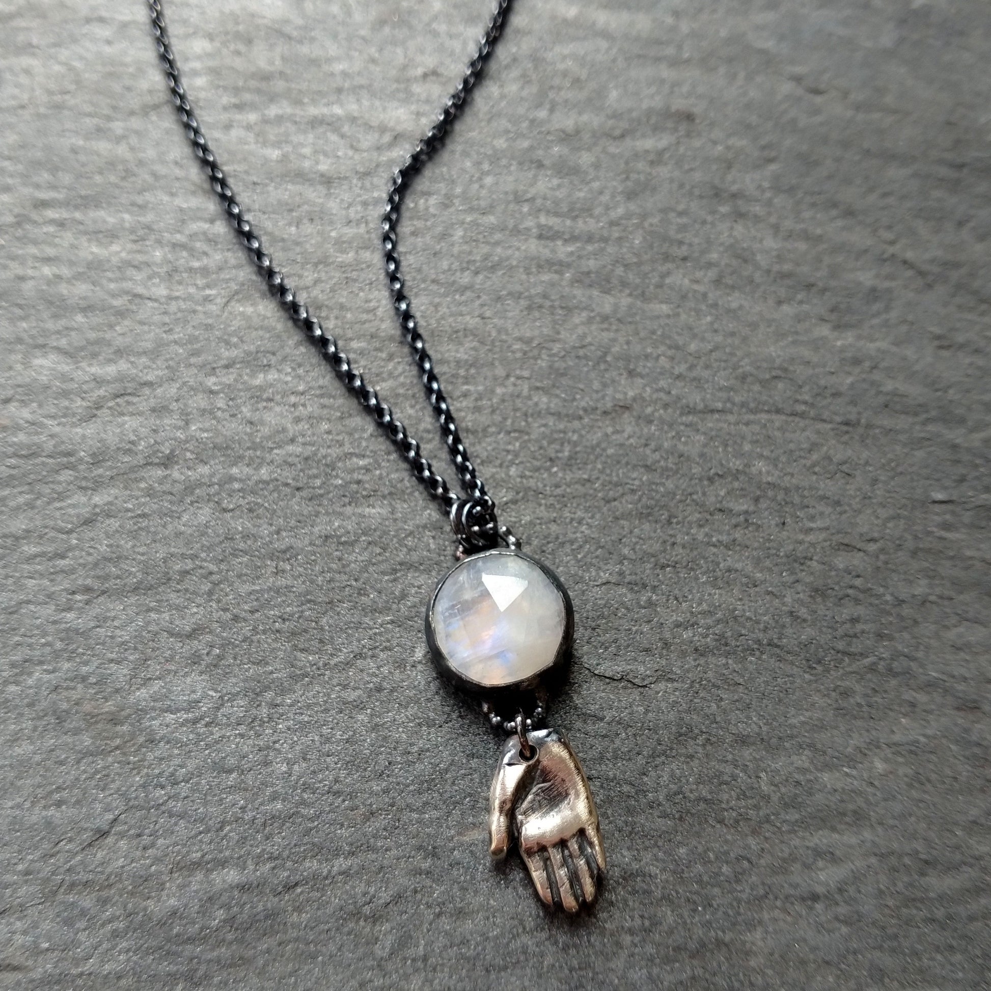 A silver necklace with a rainbow moonstone pendant and a hand talisman charm on a dark textured background, by Alice Scott