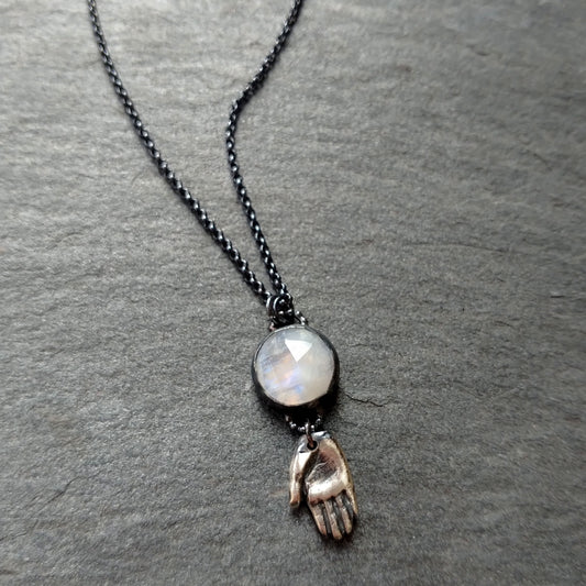 A silver necklace with a rainbow moonstone pendant and a hand talisman charm on a dark textured background, by Alice Scott