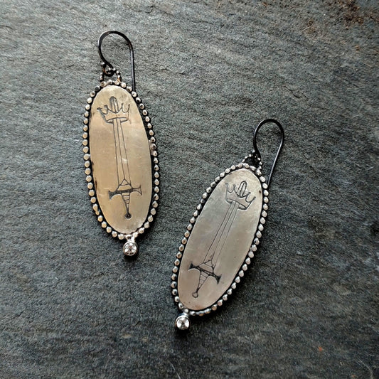 Silver Oval Earrings with sword engraving, surrounded by wilver dots and a white topaz at the bottom, by Alice Scott, displayed on a textured background.