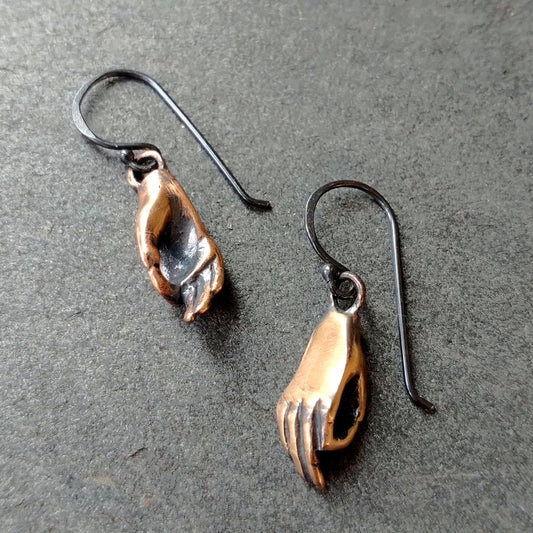 A pair of bronze earrings in the shape of hands performing a mudra, attached to oxidized silver wire hooks, set against a gray background by Alice Scott
