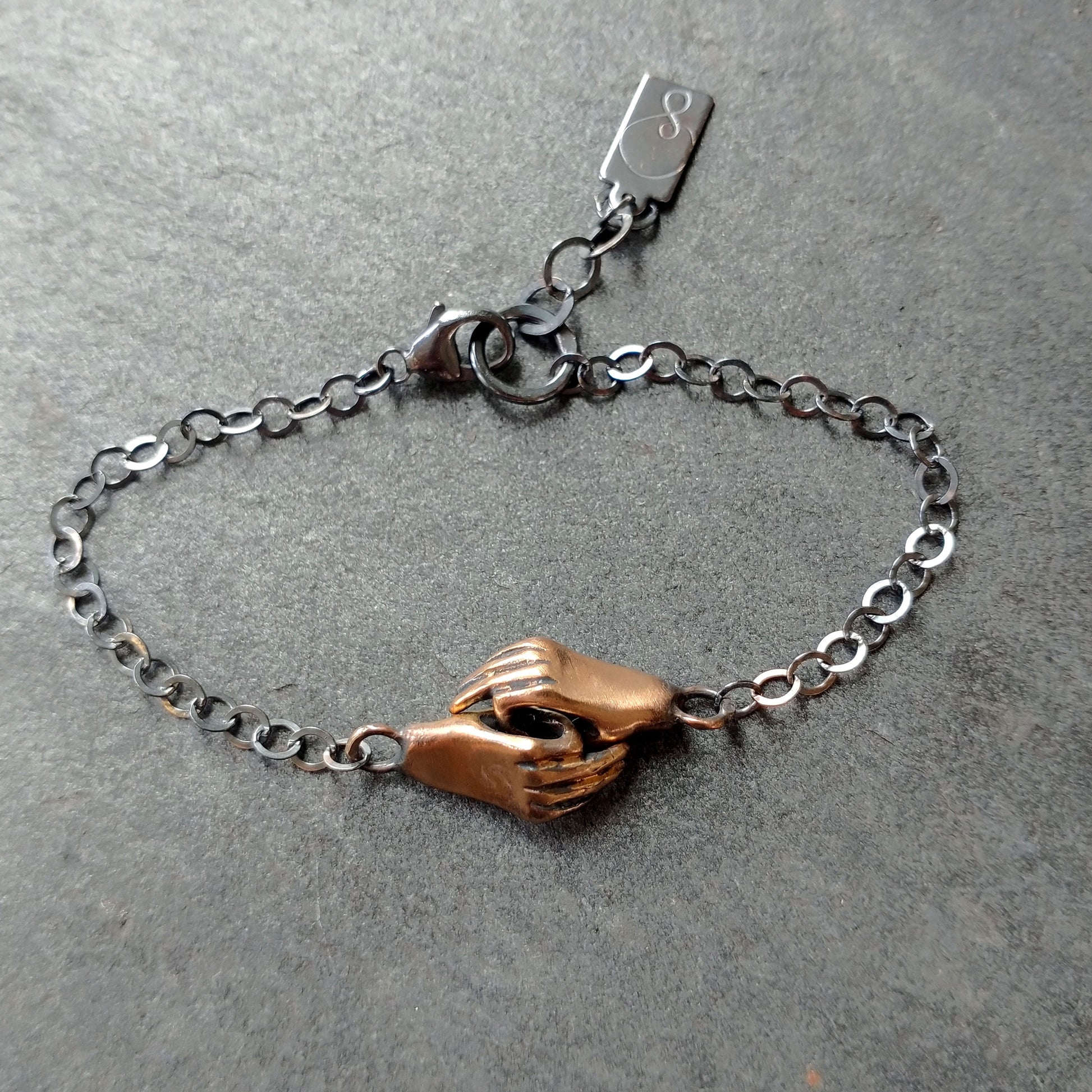 A bronze bracelet featuring a pair of hands holding each other, linked together, with a silver chain and black patina by Alice Scott on grey background