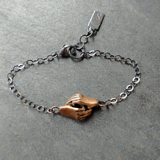 A bronze bracelet featuring a pair of hands holding each other, linked together, with a silver chain and black patina by Alice Scott on grey background
