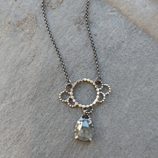Silver necklace with circlular lacy dotted pendant and rosecut aquamarine hanging below, on stone surface,  by Alice Scott
