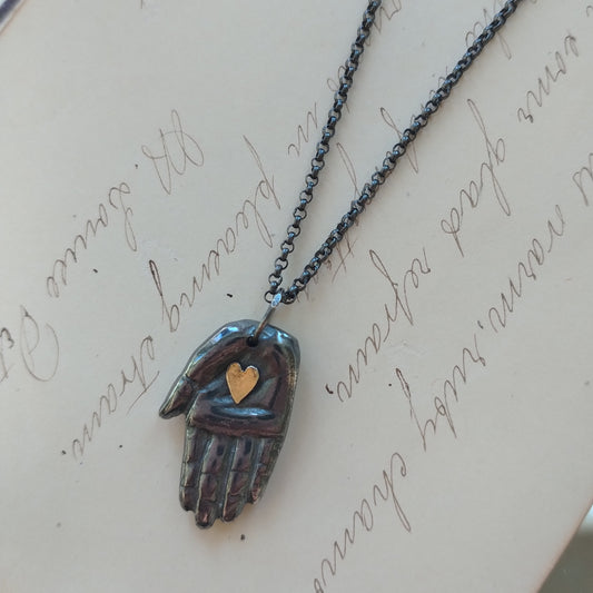 A silver necklace with a handmade patina finish featuring a hand with a gold heart pendant on a chain, displayed on a written background, by Alice Scott.